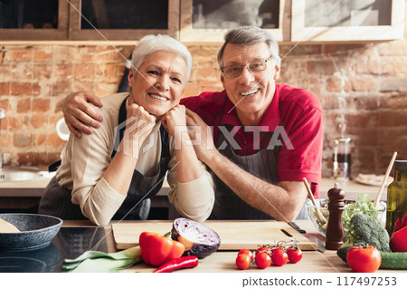 A senior couple stands side by side in their kitchen, preparing a meal. The woman is cutting a cucumber on a cutting board, while the man holds up a yellow bell pepper. They both wear aprons 117497253