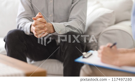 A man sits on a couch with his hands clasped, speaking with a therapist who takes notes on a clipboard during a session in a cozy living room setting, cropped 117497280