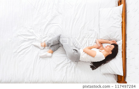 A woman is lying on a bed covered with crisp white sheets. She appears relaxed, with her eyes closed and hands resting by her side. The room is bright and minimalistic, top view 117497284