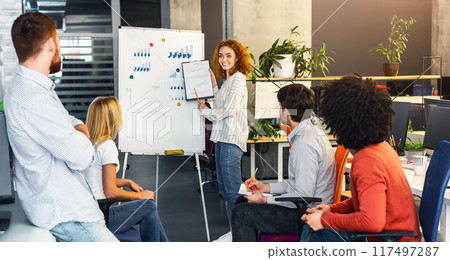 A confident young woman stands presenting a chart to a group of attentive colleagues during a collaborative meeting in a contemporary workspace A confident young woman stands presenting a chart to a group of attentive colleagues during a collaborative meeting in a contemporary workspace 117497287