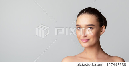 A young woman is smiling while standing in front of a plain gray background. Her expression is cheerful and relaxed as she looks directly at the camera, panorama with copy space A young woman is smiling while standing in front of a plain gray background. Her expression is cheerful and relaxed as she looks directly at the camera, panorama with copy space 117497288
