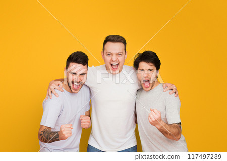 Three young men are standing together against a vibrant yellow background, arms around shoulders, with expressions of excitement and triumph Three young men are standing together against a vibrant yellow background, arms around shoulders, with expressions of excitement and triumph 117497289