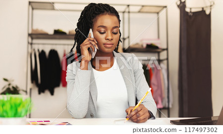 African American woman is seated at a desk, engaged in a phone conversation. She is talking on a cell phone, looking focused and attentive. The background shows office supplies and a computer. African American woman is seated at a desk, engaged in a phone conversation. She is talking on a cell phone, looking focused and attentive. The background shows office supplies and a computer. 117497302