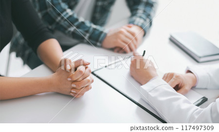 This image shows a close up of the hands of a patient and a doctor during a medical consultation. The patient has their hands clasped together on the table in front of them This image shows a close up of the hands of a patient and a doctor during a medical consultation. The patient has their hands clasped together on the table in front of them 117497309
