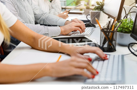 Cropped of employees seated side by side at a contemporary office desk, focused on their work on laptops. They appear to be engaged in a collaborative work environment during the day 117497311