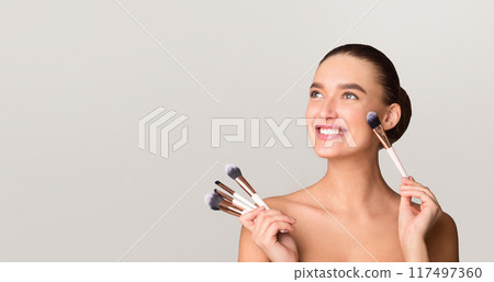 A woman smiling while holding a makeup brush in her hand. She appears happy and engaged in the act of applying makeup, empty space A woman smiling while holding a makeup brush in her hand. She appears happy and engaged in the act of applying makeup, empty space 117497360