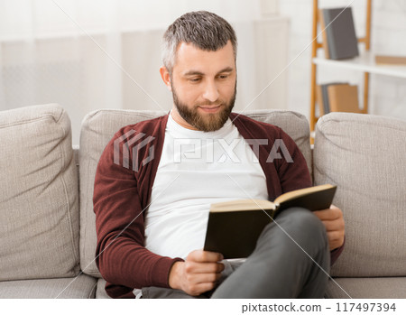 Bearded man is seated comfortably on a couch, engrossed in a book he is reading. The room is quiet as he flips through the pages, his focus solely on the words in front of him. 117497394