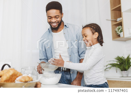 Black little girl sieving flour for pastry dough under lovely dad supervision, cooking together at kitchen, copy space 117497401
