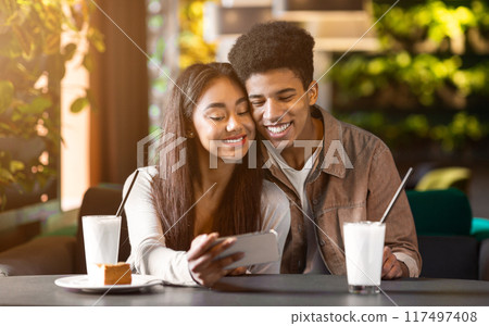 A happy African American couple sits at a cafe table, enjoying an afternoon together. They smile warmly as they take a selfie with a smartphone. A happy African American couple sits at a cafe table, enjoying an afternoon together. They smile warmly as they take a selfie with a smartphone. 117497408