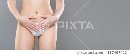 A close-up of a woman gently placing her hands on her lower abdomen, dressed in white underwear, standing against a plain grey backdrop, panorama with copy space A close-up of a woman gently placing her hands on her lower abdomen, dressed in white underwear, standing against a plain grey backdrop, panorama with copy space 117497412