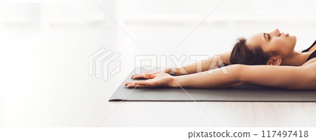 A woman is lying peacefully on a yoga mat with her arms stretched above her head, enjoying a moment of calm and relaxation in a bright, minimalistic indoor studio, panorama with copy space 117497418