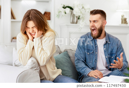 A man and woman seated on a sofa in a living room setting. 117497430