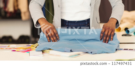 A fashion designer black woman stands at a bright work table, meticulously folding a piece of blue fabric with precision. The workstation is scattered with various sewing accessories, cropped 117497431