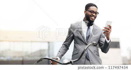 A professional black man is on his bicycle, using his phone in an urban area. The atmosphere is dynamic and contemporary, showcasing a modern approach to work and transportation. A professional black man is on his bicycle, using his phone in an urban area. The atmosphere is dynamic and contemporary, showcasing a modern approach to work and transportation. 117497434