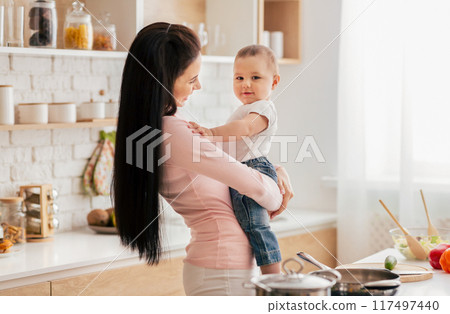 A woman stands in a kitchen, holding a baby in her arms. They appear to be bonding as they share a moment together in a typical household setting. A woman stands in a kitchen, holding a baby in her arms. They appear to be bonding as they share a moment together in a typical household setting. 117497440