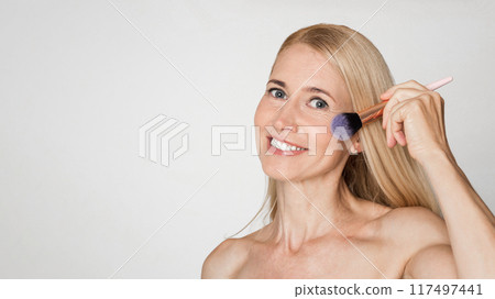 A blonde woman with a white background applies blush to her cheek using a brush. She smiles while looking at the camera. A blonde woman with a white background applies blush to her cheek using a brush. She smiles while looking at the camera. 117497441