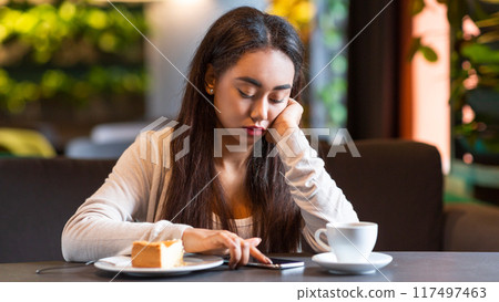 African American young girl sits alone at a cozy cafe table in the afternoon, browsing her smartphone. A slice of cake and a cup of coffee are in front of her. 117497463