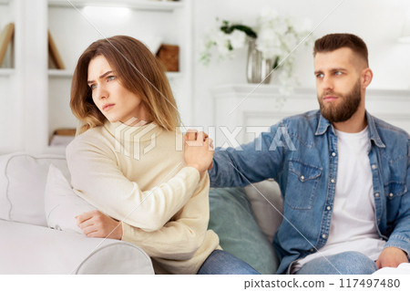 A man and a woman are seated on a modern couch in a living room setting. 117497480
