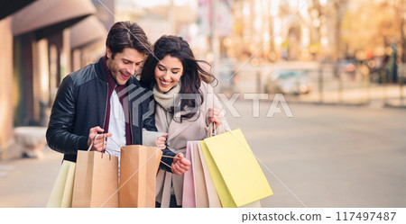 A man and woman, holding shopping bags, walk down a bustling city street on a sunny day. A man and woman, holding shopping bags, walk down a bustling city street on a sunny day. 117497487
