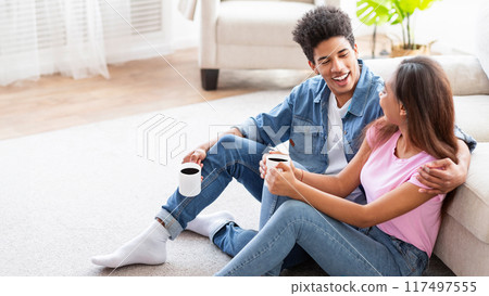 African American teen couple sits on the floor of their living room, enjoying coffee and conversation, both smiling and looking at each other, seemingly happy and relaxed. 117497555