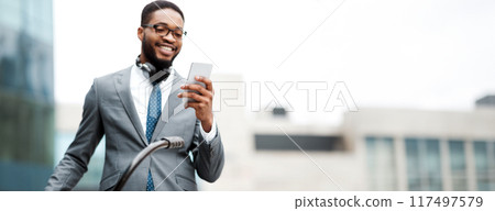 An African American businessman with his bicycle is using his phone. The mood is relaxed yet professional, capturing a modern lifestyle in an outdoor setting. 117497579