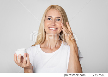 A woman with long blonde hair smiles as she applies cream to her face. She is wearing a plain white t-shirt and is standing in front of a simple grey backdrop. 117497580