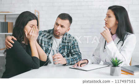 A young woman sits at a table with her partner and a doctor. The woman is crying and covering her face with her hands, while her partner looks concerned and holds a glass of water. A young woman sits at a table with her partner and a doctor. The woman is crying and covering her face with her hands, while her partner looks concerned and holds a glass of water. 117497581