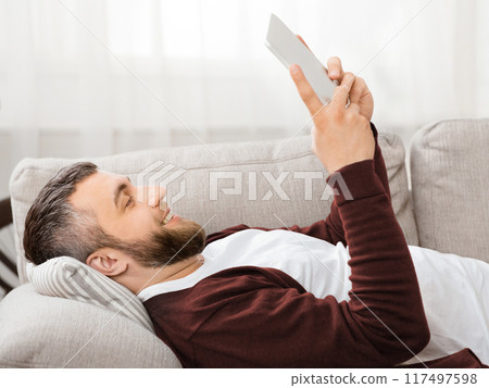 A bearded man is lying on a comfortable couch, using a tablet. The living room is well-lit with soft natural daylight coming in through the curtains, creating a serene and relaxing atmosphere. A bearded man is lying on a comfortable couch, using a tablet. The living room is well-lit with soft natural daylight coming in through the curtains, creating a serene and relaxing atmosphere. 117497598