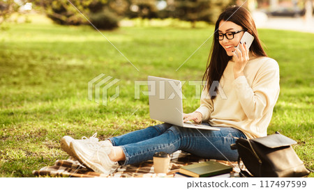 A young Asian woman is sitting on a plaid blanket in a park on a sunny afternoon, multitasking by using her laptop and talking on her phone, relaxed and focused, enjoying the outdoor environment A young Asian woman is sitting on a plaid blanket in a park on a sunny afternoon, multitasking by using her laptop and talking on her phone, relaxed and focused, enjoying the outdoor environment 117497599