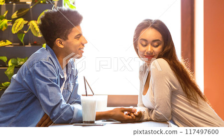 A young African American couple is sharing a moment of affection while enjoying smoothies at a cozy cafe, seated at a table, smiling and holding hands, with sunlight streaming through the window. 117497600