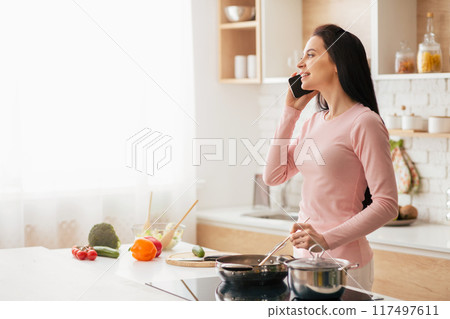 A woman standing in a kitchen area, holding a cell phone to her ear and engaged in a conversation. She is wearing casual clothing and appears focused on the call. A woman standing in a kitchen area, holding a cell phone to her ear and engaged in a conversation. She is wearing casual clothing and appears focused on the call. 117497611