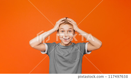 Surprised excited little boy holding hands on head, looking at camera with amazement, orange studio background Surprised excited little boy holding hands on head, looking at camera with amazement, orange studio background 117497638