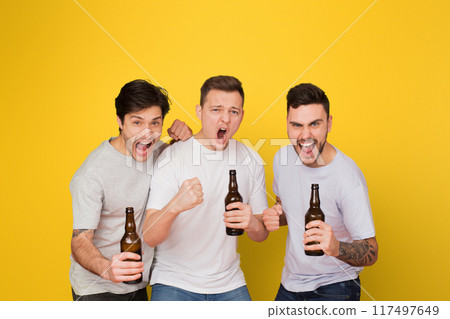 Three enthusiastic young men holding beer bottles cheer and shout together against a vivid yellow background, portraying friendship and celebration Three enthusiastic young men holding beer bottles cheer and shout together against a vivid yellow background, portraying friendship and celebration 117497649