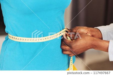 A tailor black woman is meticulously measuring the waist of a client, who is donned in a striking blue dress. The tailors hands are carefully adjusting a yellow measuring tape, cropped 117497667