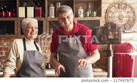 A senior couple films a cooking video in their home kitchen. The man is holding a red bell pepper and smiling at the camera, while the woman is standing behind him and smiling. 117497673