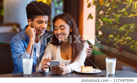 A young African American couple is seated in a cozy cafe during the afternoon, sharing earbuds and smiling as they watch something on a smartphone. They seem relaxed, enjoying each others company. 117497694