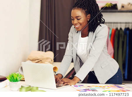 A smiling African American woman fashion designer strikes a pose with a measuring tape in her workshop, garments all around, using laptop A smiling African American woman fashion designer strikes a pose with a measuring tape in her workshop, garments all around, using laptop 117497723