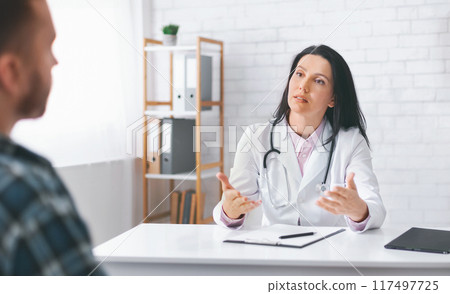 Woman doctor in a white coat and stethoscope is sitting at a desk in an office, explaining medical information to a patient who is listening attentively. The doctor has a serious expression Woman doctor in a white coat and stethoscope is sitting at a desk in an office, explaining medical information to a patient who is listening attentively. The doctor has a serious expression 117497725