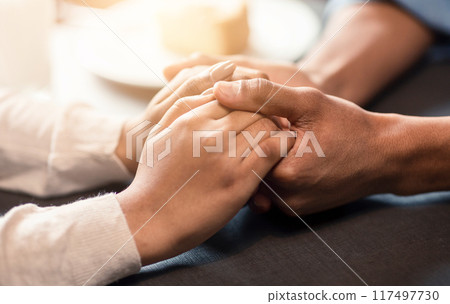 A guy and a girl are seated at a table, holding hands. The guys hand is slightly larger than the girls. Both appear relaxed and engaged in conversation, cropped 117497730