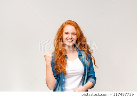 A radiant young woman with flowing red hair is captured mid-celebration, her fist pumped in victory and a beaming smile across her face, signaling triumph or good news A radiant young woman with flowing red hair is captured mid-celebration, her fist pumped in victory and a beaming smile across her face, signaling triumph or good news 117497739