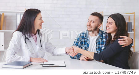 A couple sits in an office and shakes hands with a lady doctor wearing a white coat. They are all smiling and appear to be having a positive interaction 117497749