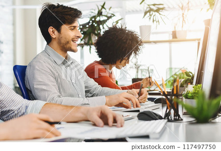 Multiethnic men are seated at a desk, engrossed in their work on computers. Each person is focused on their screen, typing and clicking away, with papers and notebooks scattered around them. 117497750