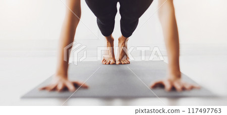 Unrecognizable woman is seen in a plank position on a yoga mat indoors. The image focuses on the legs and hands, suggesting a yoga exercise in a well-lit room during the day. Unrecognizable woman is seen in a plank position on a yoga mat indoors. The image focuses on the legs and hands, suggesting a yoga exercise in a well-lit room during the day. 117497763