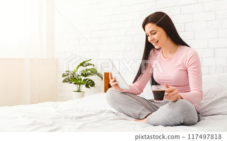 A cheerful woman sits on her bed holding a tablet and a cup of coffee, enjoying a peaceful morning in her cozy bedroom. She is dressed in comfortable pajamas with a smile on her face A cheerful woman sits on her bed holding a tablet and a cup of coffee, enjoying a peaceful morning in her cozy bedroom. She is dressed in comfortable pajamas with a smile on her face 117497818