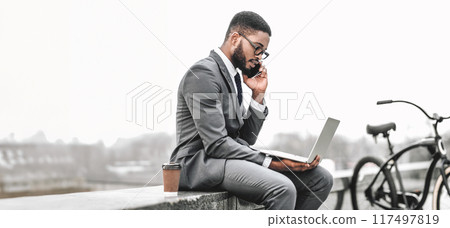 African American Businessman in a suit talking on a phone while working on a laptop outdoors with bicycle by him, panorama with copy space 117497819