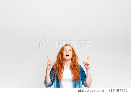 A young woman with vibrant red hair stands in front of a white backdrop, looking upwards with a look of surprise and excitement as she points at something unseen above her head, copy space 117497821