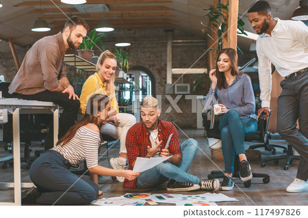 Diverse group of individuals sitting on the floor inside an office setting, engaged in a meeting or discussion. Diverse group of individuals sitting on the floor inside an office setting, engaged in a meeting or discussion. 117497826