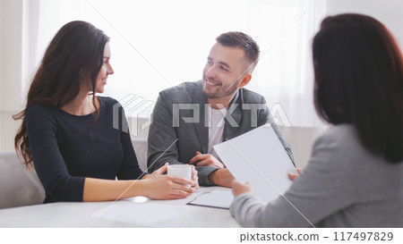 Couple sits at a table in an office, looking at documents with a financial advisor. Advisor holds up a sheet of paper while the couple looks attentive, suggesting they are in the midst of discussion 117497829