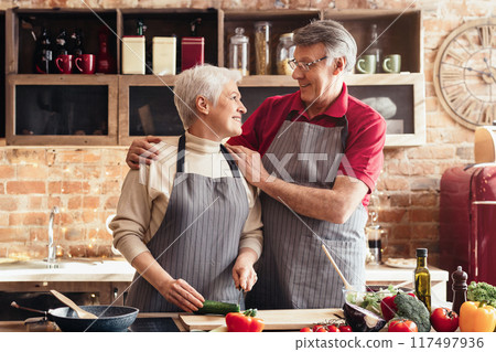 A woman is chopping a cucumber on a wooden cutting board in a kitchen, while a man in the background prepares a salad. The kitchen is bright and sunny, and the scene is one of peaceful domesticity. 117497936