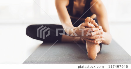 A woman is deeply engaged in her yoga practice, performing a seated forward bend stretch on a mat. The studio is filled with natural light, cropped 117497946